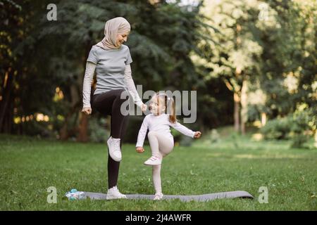 Yoga- und Stretching-Übungen von Mama mit Kind im Freien. Sympathische junge muslimische Mutter und ihre niedliche Tochter, die im Park trainieren, auf einer Yogamatte stehen und hohe Trittübungen machen Stockfoto