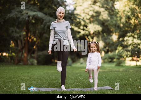 Yoga- und Stretching-Übungen von Mama mit Kind im Freien. Sympathische junge muslimische Mutter und ihre niedliche Tochter, die im Park trainieren, auf einer Yogamatte stehen und hohe Trittübungen machen Stockfoto