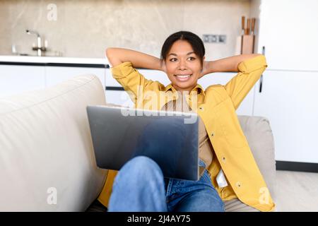 Die charmante, glückliche Afroamerikanerin ruht von der Online-Arbeit auf dem Laptop, die lächelnde Freiberuflerin lehnt sich mit den Händen hinter dem Kopf zurück und macht eine Pause auf der Couch. Remote-Work-Konzept Stockfoto