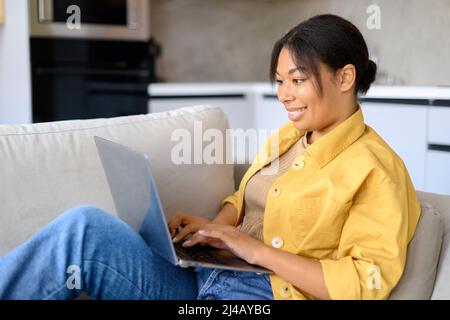 Entspannte junge afroamerikanische Freiberuflerin sitzt auf der Couch und schaut sich Filmserien auf dem Laptop an oder arbeitet aus der Ferne, tippt E-Mails, Seitenansicht Stockfoto