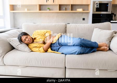 Junge afroamerikanische Frau, die an Menstruationsschmerzen leidet, sich Magenbeschwerden anfühlt, den Bauch hält, während der Periode Bauchkrämpfe und Kopfschmerzen erlebt, auf der Couch liegend Stockfoto