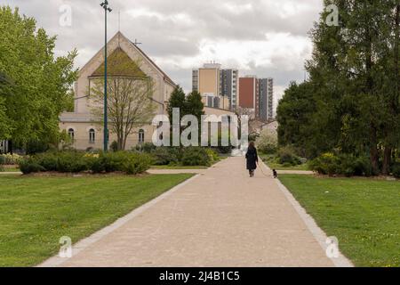 Blick auf die Ausrichtung von grünen Bäumen entlang eines Weges mit einer Fußgängerin, einem Hund, Gebäuden und einer Kirche dahinter Stockfoto