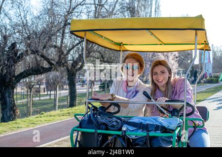 Mutter und Tochter haben Spaß beim Fahren eines überdachten Tandemfahrrads oder eines Vierrads mit einem Lenkrad auf einer Promenade. Stockfoto