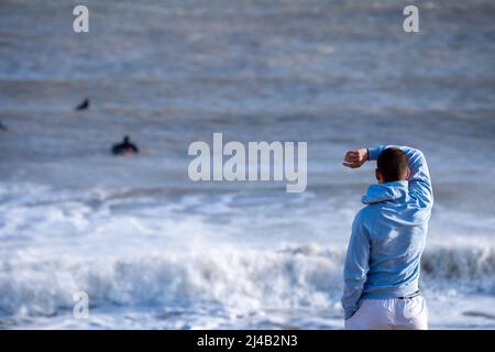 Rückansicht eines jungen Mannes am Strand von Rivedoux, der Surfer beim Wellenreiten beobachtet, Ile de Ré, Re Island, Frankreich. Stockfoto