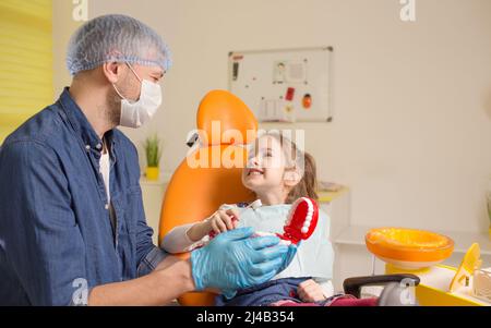 Ein männlicher Zahnarzt lehrt ein kleines Mädchen, wie sie ihre Zähne mit einer Zahnbürste richtig putzen kann. Das Konzept der Mundpflege. Stockfoto