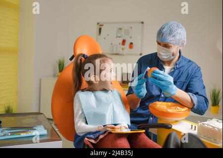 Ein männlicher Zahnarzt lehrt ein kleines Mädchen, wie sie ihre Zähne mit einer Zahnbürste richtig putzen kann. Das Konzept der Mundpflege. Stockfoto