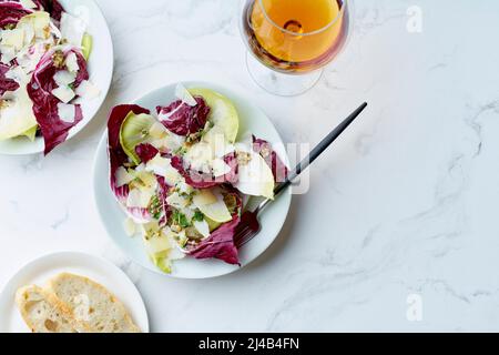 Salat mit Radicchio, Chicorée und Parmesan serviert mit einem Glas Orangenwein auf weißem Hintergrund Stockfoto