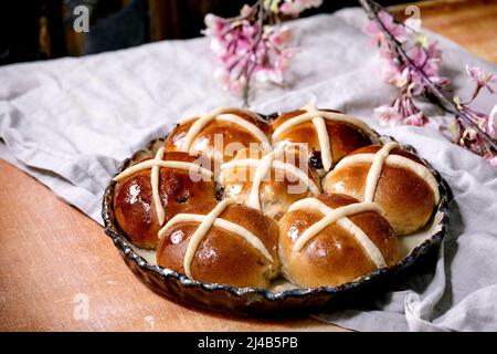 Hausgemachte Ostern traditionelle heiße Kreuz Brötchen auf Keramik-Gericht mit Blüten über orange Stein Tisch mit Leinentuch. Osterferien Backen Stockfoto
