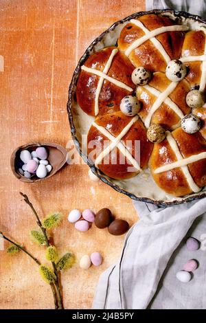 Hausgemachte Ostern traditionelle heiße Kreuz Brötchen in Keramikschale mit Blütenweiden Zweige und Schokolade Eier Süßigkeiten auf Orangenstein Hintergrund. Flach Stockfoto