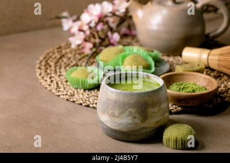 Asiatischer Reis Dessert süßer grüner Matcha-Mochi mit einer Tasse aufgeschäumtem Matcha-Tee und grünem Matcha-Pulver in Keramikplatte auf braunem Tisch. Teekannen und Frühling Stockfoto