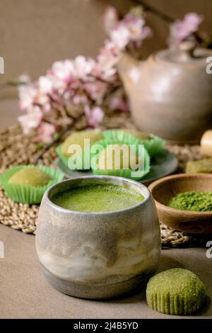 Asiatischer Reis Dessert süßer grüner Matcha-Mochi mit einer Tasse aufgeschäumtem Matcha-Tee und grünem Matcha-Pulver in Keramikplatte auf braunem Tisch. Teekannen und Frühling Stockfoto