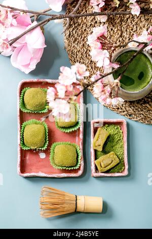 Asiatische traditionelle Reis Dessert süßen grünen Matcha-Mochi in rosa Keramik-Schale mit Tasse aufgeschäumter Matcha-Tee auf blauem Tisch mit Frühling rosa Blüten. Fl Stockfoto