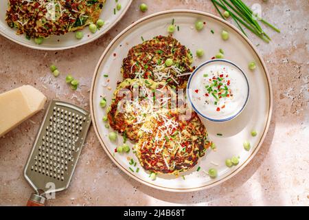 Zucchini-Pfannkuchen, Erbsen, Schnittlauch, geriebener Parmesan, Käsemühle, Topf mit würziger Creme, auf weißem Teller Stockfoto