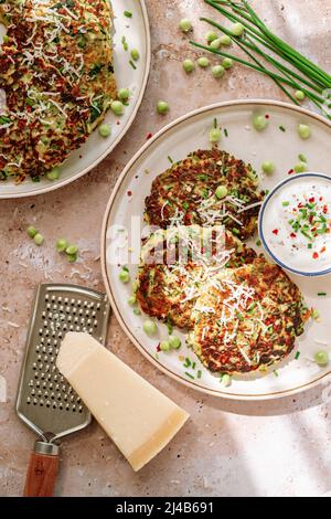 Zucchini-Pfannkuchen, Erbsen, Schnittlauch, geriebener Parmesan, Käsemühle, Topf mit würziger Creme, auf weißem Teller Stockfoto