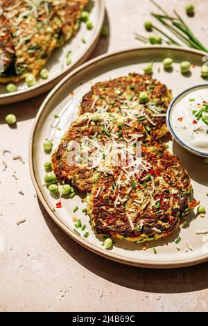 Zucchini-Pfannkuchen, Erbsen, Schnittlauch, geriebener Parmesan, Topf mit würziger Creme, auf weißem Teller Stockfoto