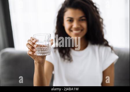 Gesundes Lifestyle-Konzept. Lächelnde, attraktive afroamerikanische Frau sitzt auf dem Sofa im Wohnzimmer und hält ein Glas Wasser in der Hand. Schöne weibliche ist folgen gesunden Lebensstil Stockfoto