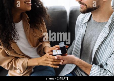 Ein Heiratsangebot. Multirassisches Paar verliebt zu Hause auf der Couch im Wohnzimmer. Der kaukasische Mann gibt seiner geliebten afroamerikanischen Frau einen Verlobungsring, den sie gerne annimmt Stockfoto