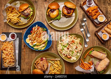 Set von Fast-Food-Gerichten auf Holztisch mit verschiedenen Burgern, Sandwiches, Süßkartoffeln, Pommes frites, tequeños, gebratenen Hähnchenstreifen und caesar-Salat, Chicke Stockfoto