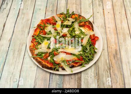 Eine leckere Pizza mit Parmesan-Käsesavertäfelchen, Rukula im Überfluss, Pomodoro-Sauce und gehackten Kirschtomaten mit spanischem Olivenöl auf einem Holztisch Stockfoto