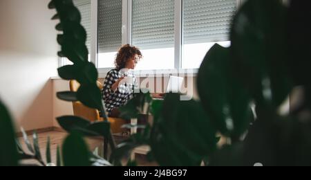 Fernansicht einer Frau, die einen Videoanruf mit Freunden über einen Laptop-Computer macht, sich zu Hause auf den Stuhl setzt und eine Tasse Kaffee trinkt. Unternehmen Stockfoto