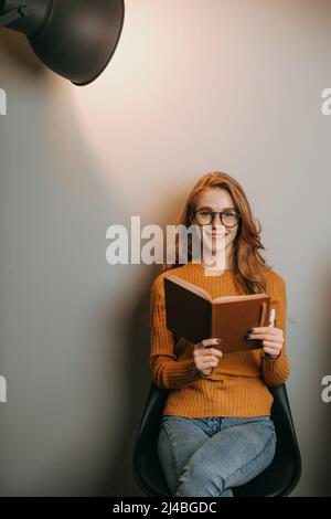 Eine rotköpfige Frau sitzt auf einem Stuhl über einer weißen Wand und liest ein Buch, das im Scheinwerferlicht posiert. Business, Bildungskonzept. Modernes kreatives Geschäft Stockfoto