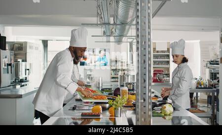 afroamerikanische kochen schneiden Paprika und Zugabe in der Pfanne, Kochen Gemüse Küche Rezept. Koch schneidet Zutaten gleichmäßig auf dem Schneidebrett und bereitet ein professionelles Gourmetgericht zu. Stockfoto