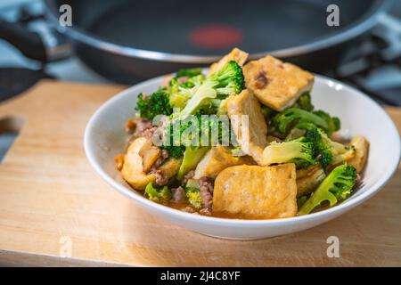 Gebratener Brokkoli und gebratener weicher Tofu, gesundes asiatisches hausgemachtes Menü, Essen auf weißem Teller, der in einer Küche auf Holzschneidebrett platziert wird, Nahaufnahme Th Stockfoto