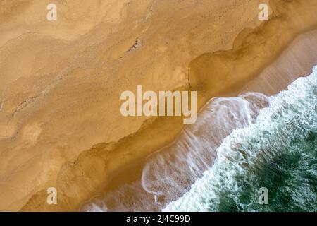 Drone-Ansicht von oben nach unten von weißgedeckten Wellen, die an einem leeren goldenen Sandstrand zusammenbrechen Stockfoto