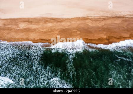 Drone-Ansicht von oben nach unten von weißgedeckten Wellen, die an einem leeren goldenen Sandstrand zusammenbrechen Stockfoto