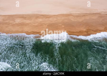 Drone-Ansicht von oben nach unten von weißgedeckten Wellen, die an einem leeren goldenen Sandstrand zusammenbrechen Stockfoto