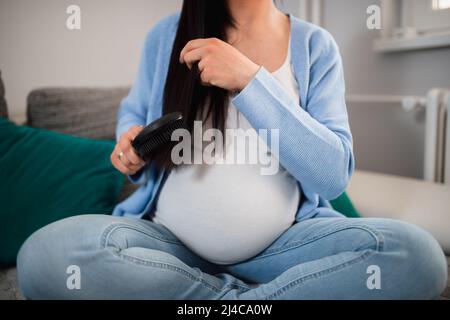 Eine unkenntlich werdende, schwanger kaukasische Frau kämmt ihr langes, poliertes Haar mit einer schwarzen Haarbürste. Sie sitzt mit gekreuzten Beinen auf dem Sofa. Stockfoto