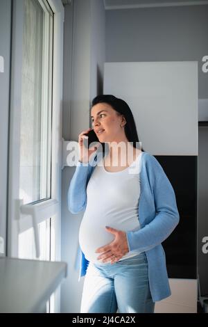Eine gut aussehende kaukasische Schwangerin ist in der Wohnung. Eine wunderschöne Frau spricht auf ihrem Handy, während sie durch die Tür schaut. Stockfoto