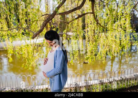 Eine junge kaukasische Frau in fortgeschrittener Schwangerschaft kuschelt ihren Bauch mit beiden Händen und schaut ihn an. Eine schwarzhaarige Schwangere steht neben dem See. Stockfoto