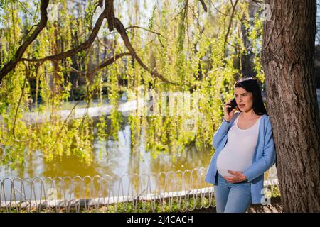 Eine gut aussehende, schwarzhaarige kaukasische, kindertragende Schwangere telefoniert im grünen Park. Sie lehnt sich an einen Baum in der Nähe des Sees und schaut nachdenklich. Stockfoto