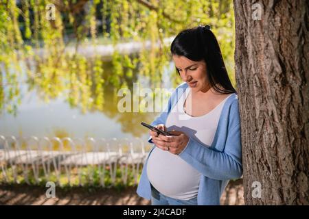 Eine kaukasische Schwangere lehnt sich gut gelaunt an den Baum im Park in der Nähe des Sees. Eine kindgebärende Frau schaut zufrieden auf den Telefonbildschirm. Stockfoto