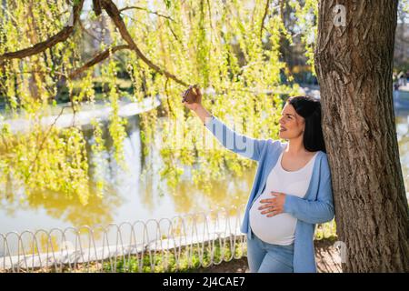 Eine attraktive Schwangere macht Selfies mit ihrem Smartphone, während sie sich in einem Park an den Baum lehnt. Ihre linke Hand liegt auf dem Bauch. Stockfoto