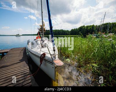 An einem sonnigen Sommertag wurde ein Segelboot in der Nähe eines alten hölzernen Piers an einem Seeufer festgemacht Stockfoto