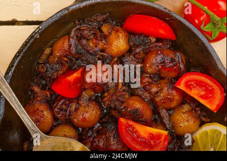 Baby Tintenfisch über rustikalen Holztisch auf eisernen Pfanne mit Tomaten und Zwiebeln gebraten Stockfoto