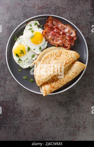 Englisches Frühstück mit Speck, Eiern und Haferflocken Haferpfannkuchen aus der Nähe auf einem Teller auf dem Tisch. Vertikale Draufsicht von oben Stockfoto