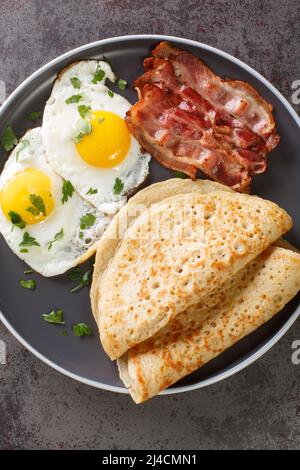 Traditionelle englische Staffordshire Haferpfannkuchen, Speck und Spiegeleier in Nahaufnahme auf einem Teller auf dem Tisch. Vertikale Draufsicht von oben Stockfoto