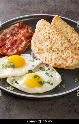 Traditionelle englische Staffordshire Haferpfannkuchen, Speck und Spiegeleier in Nahaufnahme auf einem Teller auf dem Tisch. Vertikal Stockfoto