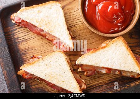 Bacon butty ist ein britisches Sandwich, das aus knusprigem Speck, Butter und Sauce in der Nähe auf dem Holztablett auf dem Tisch besteht. Horizontale Draufsicht von oben Stockfoto