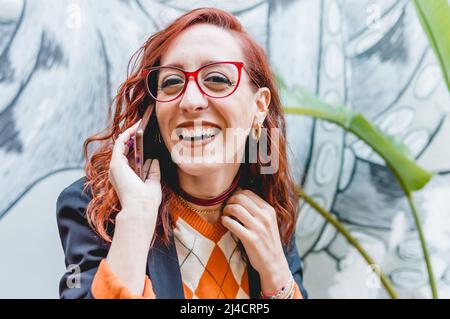 Junge kaukasische Rotschopf-Geschäftsfrau mit Brille und blauem Blazer, glücklich lächelnd im Freien telefonieren, Kamera, Technologiekonzept, Stockfoto