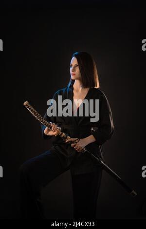 Junge schöne Frau in schwarzem Kimono mit Katana auf schwarzem Hintergrund. Stockfoto