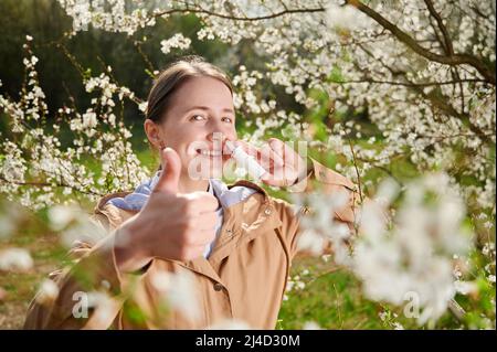 Allergische Frau, die im Frühling an einer saisonalen Allergie leidet und im Frühling im blühenden Garten posiert. Glückliche Frau mit nasalen Tropfen, zeigt Daumen hoch zwischen blühenden Bäumen. Frühjahrsallergiekonzept Stockfoto