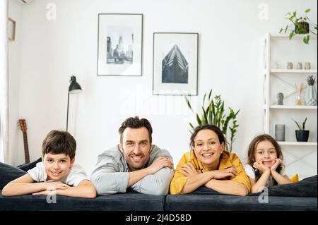 Fröhliche, multirassische vierköpfige Familie, die auf dem Sofa im gemütlichen Wohnzimmer liegt, Eltern und Kinder lachen. Familie, die sich am Wochenende glücklich fühlt Stockfoto