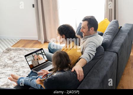 Vierköpfige Familie mit Laptop für Videoverbindung mit Großeltern oder Familie. Multirassische Mutter, Vater und zwei Kinder winken in der Webcam des Laptops, Videoanruf, virtuelles Konferenzkonzept Stockfoto