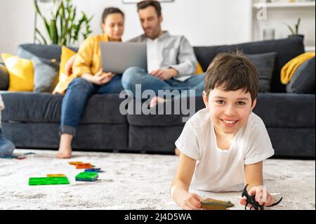Zwei Eltern sitzen auf dem Sofa im gemütlichen Wohnzimmer und schauen auf den Laptop, während ihr Sohn auf dem Boden spielt. Familie zu Hause Stockfoto