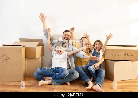Eine glückliche, multirassische Familie von vier Personen, die auf dem Boden sitzen und Kartons in einem leeren Wohnzimmer umgeben. Besitzer von Immobilien, Hypothek Stockfoto