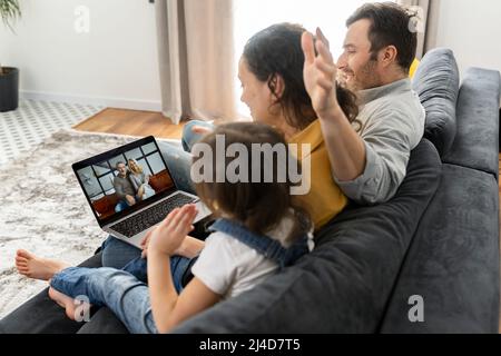 Vierköpfige Familie mit Laptop für Videoverbindung mit Großeltern oder Familie. Multirassische Mutter, Vater und zwei Kinder winken in der Webcam des Laptops, Videoanruf, virtuelles Konferenzkonzept Stockfoto